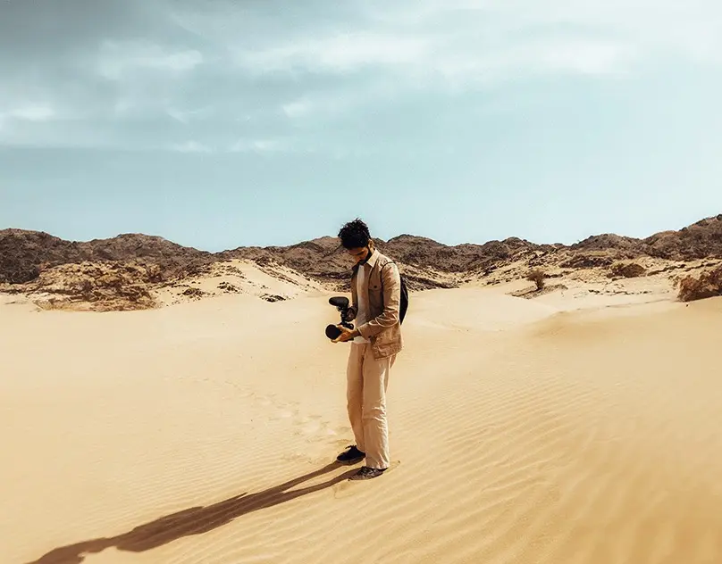 عزّام يحمل كاميرا سينمائية على كثيب رملي تحت سماء صافية